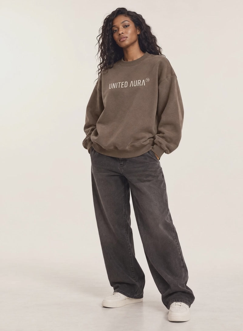 VIDEO OF A model stands against a plain light background wearing a brown crewneck sweatshirt with 'UNITED AURA' printed in white across the chest, paired with dark gray wide-leg jeans and white sneakers.